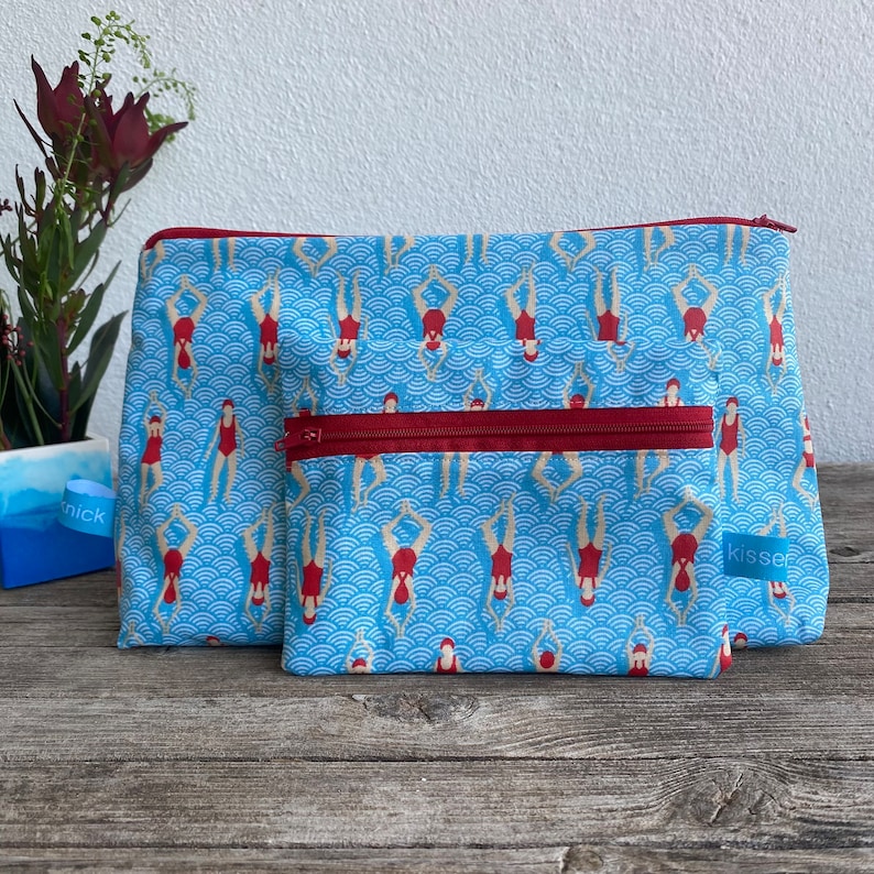 Peut inclure: Deux trousses de toilette bleues avec une fermeture &eacute;clair rouge et un motif de nageurs en maillots de bain rouges. Les sacs ont un fond bleu clair avec un motif de vagues. Les sacs sont de tailles diff&eacute;rentes et sont en tissu.