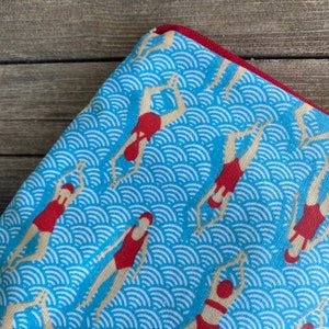 Peut inclure: Tissu bleu avec un motif r&eacute;p&eacute;t&eacute; de nageurs en maillots de bain et bonnets rouges, sur un fond de motifs blancs en forme de vagues. Le bord du tissu est bord&eacute; d'une bordure rouge.