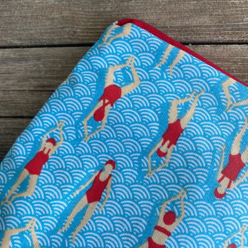 Peut inclure: Tissu bleu avec un motif r&eacute;p&eacute;t&eacute; de nageurs en maillots de bain et bonnets rouges, sur un fond de motifs blancs en forme de vagues. Le bord du tissu est bord&eacute; d'une bordure rouge.