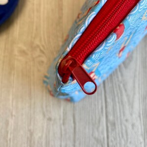 Peut inclure: Une pochette rectangulaire en tissu bleu clair avec une fermeture &eacute;clair rouge. La pochette pr&eacute;sente un motif de personnages rouges et blancs sur un fond bleu clair. La fermeture &eacute;clair rouge est ouverte, r&eacute;v&eacute;lant l'int&eacute;rieur rouge de la pochette.