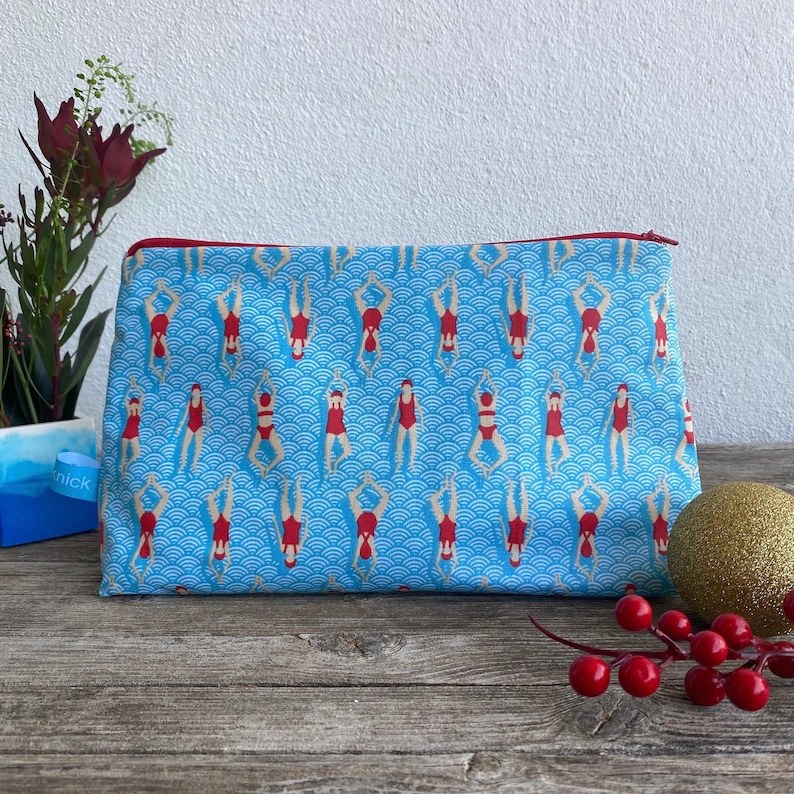 Peut inclure: Une trousse de maquillage rectangulaire avec un fond bleu orn&eacute; d'un motif de nageurs en maillots de bain rouges. Le sac a une fermeture &eacute;clair rouge et est pos&eacute; sur une surface en bois.