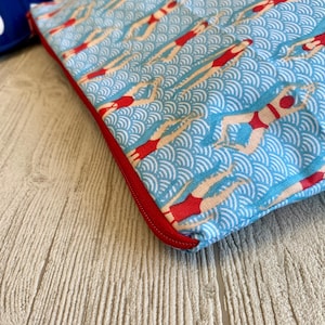 Peut inclure: Une pochette en tissu bleu avec un motif r&eacute;p&eacute;t&eacute; de nageurs en maillots de bain rouges sur un fond bleu clair ondul&eacute;. La pochette a une fermeture &eacute;clair et une bordure rouges. Th&egrave;me aquatique r&eacute;tro.