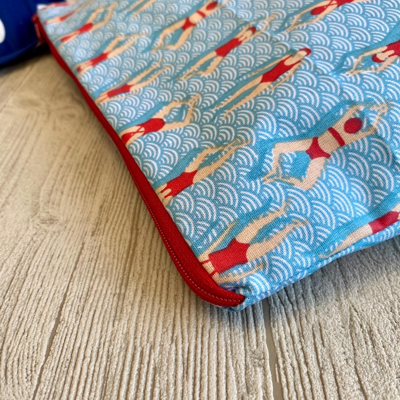 Peut inclure: Une pochette en tissu bleu avec un motif r&eacute;p&eacute;t&eacute; de nageurs en maillots de bain rouges sur un fond bleu clair ondul&eacute;. La pochette a une fermeture &eacute;clair et une bordure rouges. Th&egrave;me aquatique r&eacute;tro.