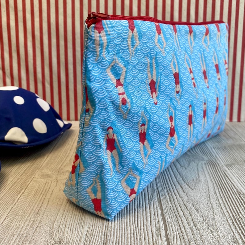 Peut inclure: Une trousse de maquillage rectangulaire avec un fond bleu orn&eacute; d'un motif de nageurs en maillots de bain rouges. Le sac a une fermeture &eacute;clair et une doublure rouges. Un article bleu et blanc &agrave; pois est visible en arri&egrave;re-plan.