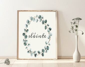 Può includere: Una stampa incorniciata con una ghirlanda di eucalipto ad acquerello nei toni del verde e del verde-blu. La parola "sláinte" è scritta in elegante corsivo all'interno della ghirlanda. La cornice è di colore marrone chiaro e lo sfondo è bianco sporco.
