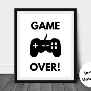 Pu&ograve; includere: Stampa con cornice nera con le parole "GAME OVER!" in grassetto e maiuscolo sopra una grafica di controller nera. Un cerchio bianco con il testo "Instant Download" si trova nell'angolo in basso a destra. La stampa &egrave; esposta su una superficie di legno scuro.