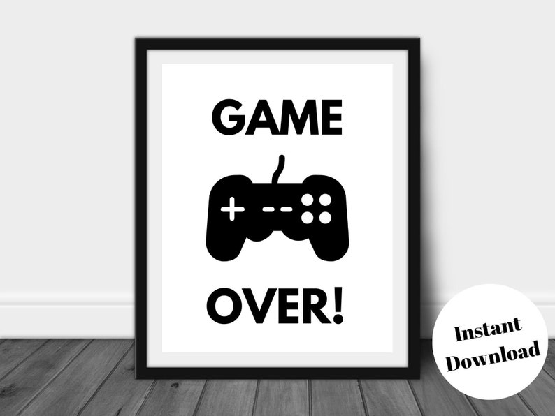 Pu&ograve; includere: Stampa con cornice nera con le parole "GAME OVER!" in grassetto e maiuscolo sopra una grafica di controller nera. Un cerchio bianco con il testo "Instant Download" si trova nell'angolo in basso a destra. La stampa &egrave; esposta su una superficie di legno scuro.
