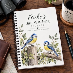 Pode incluir: Um diário de espiral branco intitulado Mike's Bird Watching Journal com um ícone de coração e o texto Notes, Sightings, Memories. A capa apresenta uma ilustração em aguarela de dois chapins-azuis num poste de madeira e um ramo.