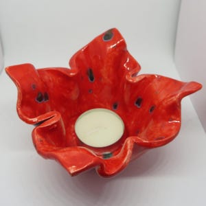 Op de afbeelding: Een rode keramische kaarsenhouder met een gegolfde rand en zwarte vlekken. Een witte theelichtkaars is in de houder geplaatst.