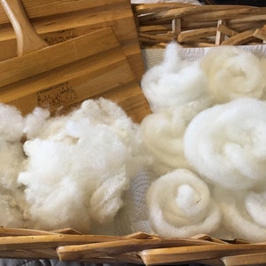 May include: A woven basket filled with fluffy white wool roving and two wooden wool carders. The carders are light brown with handles and text. The wool is arranged in loose clumps and rolled bundles, ready for fiber art projects.