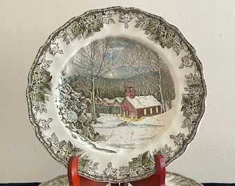 Può includere: Un set di otto piatti piani in ceramica bianca con un bordo floreale verde e marrone e una scena invernale al centro. La scena rappresenta un villaggio innevato con una scuola rossa.