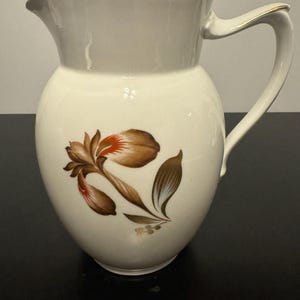 May include: A white ceramic pitcher with a gold rim and handle. The pitcher features a brown and red floral design.