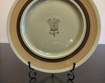 May include: A light green ceramic dinner plate with a brown and tan rim. The plate has a stylized flower design in the center.
