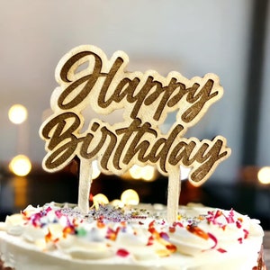 May include: A wooden cake topper with the words "Happy Birthday" in a cursive font. The topper is on top of a white cake with colorful sprinkles.