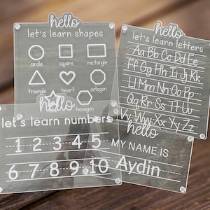 Op de afbeelding: Vier transparante, droog uitwisbare borden van acryl met witte letters. Het eerste bord zegt "hello let's learn shapes" en toont een cirkel, een vierkant, een rechthoek, een driehoek, een hart, een zeshoek en een achthoek. Het tweede bord zegt "hello let's learn letters" en toont het alfabet in kleine letters. Het derde bord zegt "hello let's learn numbers" en toont de getallen 1 tot en met 10. Het vierde bord zegt "hello my name is Aydin".