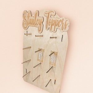 Op de afbeelding: Houten topperstand met de tekst "Stanley Toppers" bovenaan. De stand heeft 10 slots voor toppers, waarvan twee slots gevuld zijn met toppers die "Tina" en "Kim" zeggen.