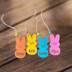 May include: Four colorful wooden bunny-shaped Easter ornaments with twine and beads. The bunnies are orange, yellow, blue, and pink. The names Oliver, Axel, Xavier, and Mya are engraved on the bunnies.