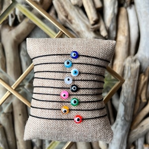 Peut inclure: Un ensemble de dix bracelets œil-de-dieu colorés sur une ficelle noire. Les bracelets sont présentés sur une surface en tissu beige.