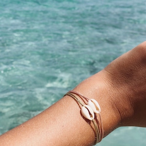 Peut inclure: Un bracelet en cordon marron et or avec deux coquillages blancs sur le poignet d'une personne. Le bracelet est noué.