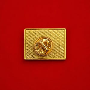 May include: A rectangular gold-tone metal pin back featuring a textured cross-hatch pattern on the surface. The center of the pin displays a circular clutch mechanism with a spring-loaded butterfly clasp for secure attachment to fabric.