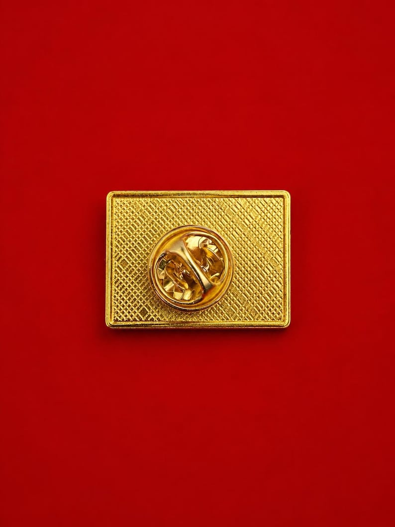 May include: A rectangular gold-tone metal pin back featuring a textured cross-hatch pattern on the surface. The center of the pin displays a circular clutch mechanism with a spring-loaded butterfly clasp for secure attachment to fabric.