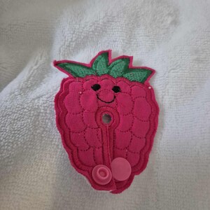 May include: A pink raspberry-shaped fabric patch with a smiling face and two pink buttons. The patch is designed for sewing onto clothing or other fabric items.