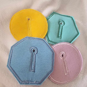 Può includere: Quattro toppe in tessuto colorate in giallo, verde acqua, azzurro e rosa, ciascuna con un design a forma di buco della serratura. Le toppe hanno un bordo cucito e sono in varie forme geometriche, tra cui un cerchio e un esagono.