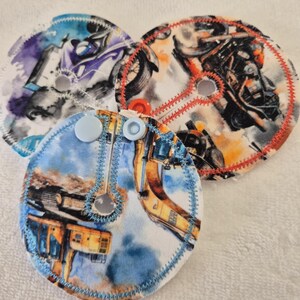 May include: Three round, reusable nursing pads with colorful designs. One pad features a motorcycle, another a construction vehicle, and the third a futuristic vehicle. Each pad has a button closure and decorative stitching around the edges.