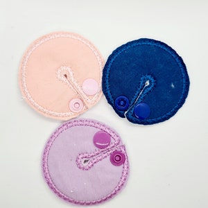 May include: Three round fabric patches with a slit and snap closure. The patches are pink, purple, and blue.