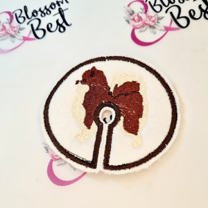 Peut inclure: Un patch brodé blanc et marron représentant un chien poméranien avec un corps marron et une queue et une poitrine blanches. Le patch est circulaire avec une bordure marron et un trou au centre.
