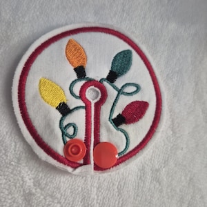 May include: Embroidered white circular patch featuring colorful Christmas lights. The design includes orange, green, yellow, and red bulbs with black details, outlined by a red border. Two orange buttons are attached at the bottom.