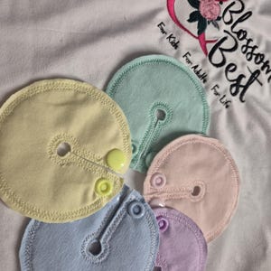 Puede incluir: Un conjunto de discos de lactancia de colores pastel en amarillo, azul, rosa, morado y verde, cada uno con cierre de botón y una abertura central. Los discos están colocados sobre una tela blanca con texto bordado y un diseño de corazón floral.