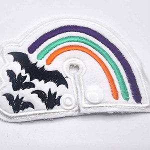 Könnte beinhalten: Ein weißer Stoff-Patch mit einem Regenbogen und Fledermäusen, die darauf gestickt sind. Der Regenbogen ist aus lila, grünem und orangefarbenem Garn gefertigt. Die Fledermäuse sind schwarz. Der Patch hat zwei weiße Druckknöpfe an den Seiten.
