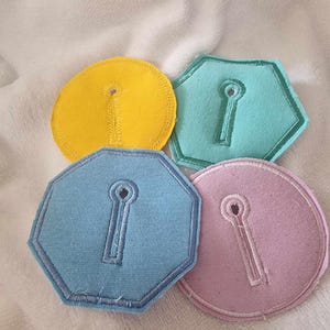 Puede incluir: Cuatro formas de ojo de cerradura de tela en diferentes colores: amarillo, azul claro, verde azulado y rosa claro. Las formas son todas de tamaños ligeramente diferentes y tienen un ojo de cerradura recortado en el centro.