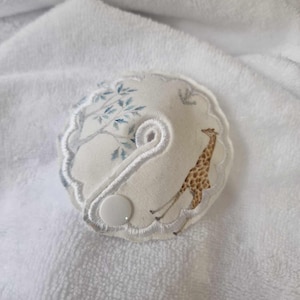 Op de afbeelding: Een witte stoffen baby spuugdoek met een witte knoop en een blauwe en bruine giraffenprint.