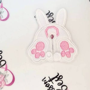May include: White embroidered bunny butt applique with pink paw pads and a pink circle detail. The applique has a white outline and two small white circles at the bottom. The background is white with pink floral accents and black script.