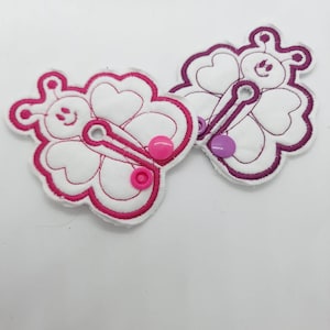 Puede incluir: Dos botones de tela blanca en forma de mariposa con bordados rosa y morado. Los botones tienen un agujero en el centro y están decorados con botones rosa y morado.
