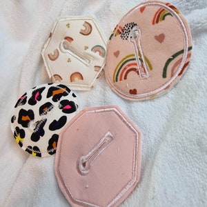 May include: Four fabric patches with different designs. Each patch has a white loop sewn on it. The patches are in various shapes, including a hexagon, a circle, and an octagon. The designs include a leopard print, a pink background with rainbows and hearts, and a white background with rainbows and small stars.