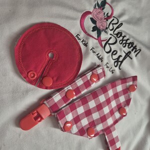 May include: A circular solid pink fabric accessory with a central hole and snap closure, alongside two pink and white gingham patterned fabric straps featuring plastic clips and snap buttons, resting on a white fabric with embroidered text Blossom Best.
