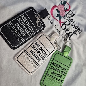 May include: Three rectangular luggage-style tags in black, white, and light green with textured surfaces. Each tag features the embroidered text MEDICAL SUPPLIES INSIDE and HANDLE WITH CARE in black or white thread, with a metal lobster clasp attached.