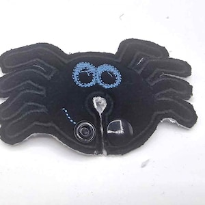 Pode incluir: Aranha de feltro preto com olhos azuis e um centro branco. A aranha tem um botão de cada lado.