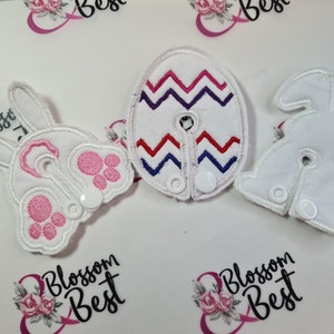 Puede incluir: Tres parches de tela blanca con diferentes diseños. Un parche presenta un conejo, otro un patrón en zigzag y el último una simple forma de huevo. Todos los parches tienen acentos rosas y azules.