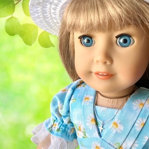 May include: A doll with blue eyes and blonde hair, wearing a blue dress with white daisy patterns and a white hat. A silver necklace with a cross pendant is visible. The doll is set against a blurred green background.