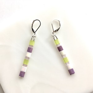 May include: A pair of dangle earrings with a geometric design. The earrings are made of purple, white, and green beads. The beads are arranged in a striped pattern.