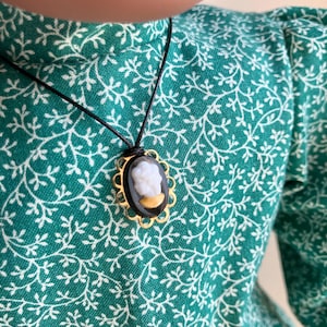May include: A gold-toned cameo pendant necklace with a black background and a white profile. The pendant is oval-shaped with a decorative gold frame. The necklace is on a green dress with a white floral pattern.