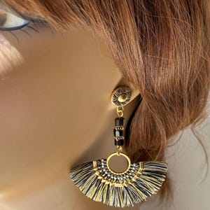 May include: Gold-toned earrings with a sunburst stud and a fan-shaped tassel design. The earrings feature black and gold tassels, black beads, and a gold ring. The earrings are a statement piece.