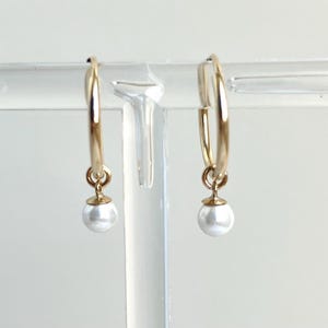 May include: A pair of gold hoop earrings with a small white pearl pendant. The earrings are displayed on a clear, horizontal stand against a white background. The hoops are circular and have a simple, elegant design.