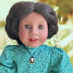 May include: A doll with dark skin, brown eyes, and dark hair styled in a bun. She wears a green dress with a white floral pattern and a cameo pendant necklace. The doll has a gold hoop earring in one ear.