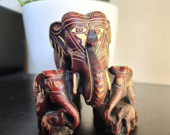 Op de afbeelding: Een handgesneden houten olifantenbeeldje met ingewikkelde details. De olifant is geschilderd in tinten rood en bruin met gouden accenten. De olifant staat op zijn vier poten met zijn slurf omhoog.