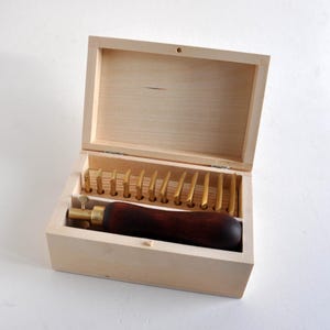 Puede incluir: Una caja de almacenamiento de madera clara que contiene un juego de doce cuchillas artesanales de metal dorado dispuestas en fila y una herramienta con mango de madera marrón oscuro y punta de latón, todo dentro del contenedor rectangular abierto.